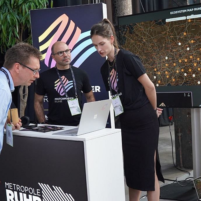 Zu sehen sind zwei Personen im Matchmaker.Ruhr T-Shirt, die einem Interessenten die Plattform erklären. Im Hintergrund ist auf einem Bildschrim das digitale Netzwerk zu sehen.