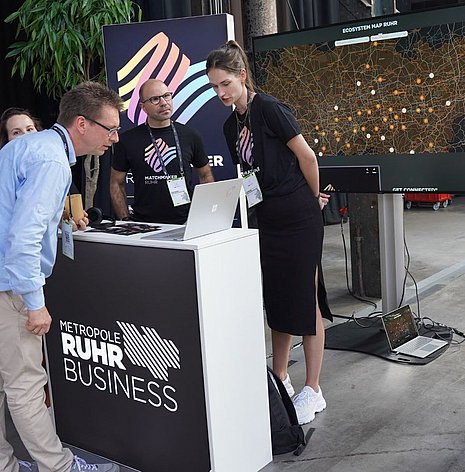 Zu sehen sind zwei Personen im Matchmaker.Ruhr T-Shirt, die einem Interessenten die Plattform erklären. Im Hintergrund ist auf einem Bildschrim das digitale Netzwerk zu sehen.