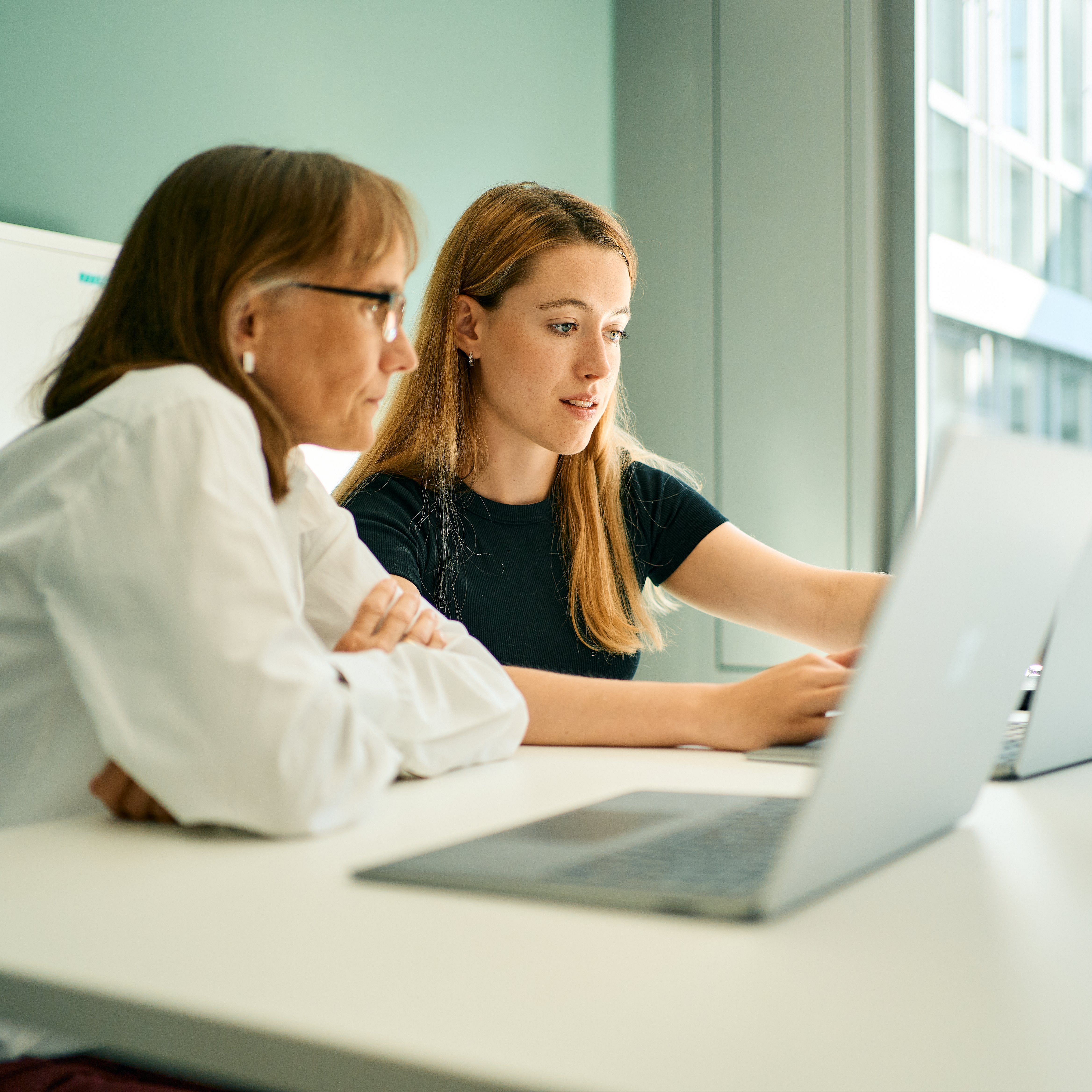 Two female employees discuss together in front of their laptops.