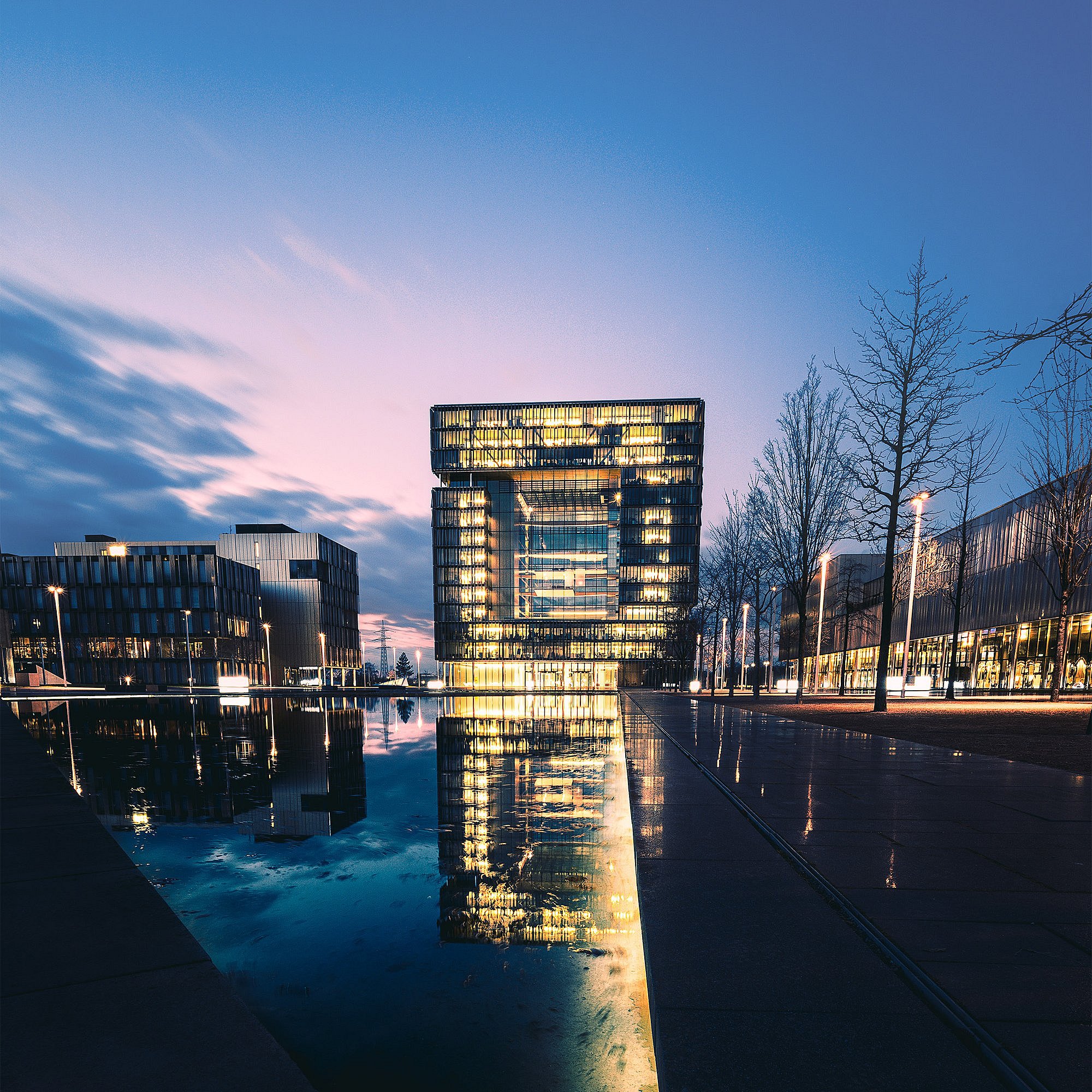 Moderne Immobilien im Ruhrgebiet wie das hier abgebildete thyssenkrupp Quartier in Essen. Zu sehen ist das beleuchtete Bürogebäude bei Abendhimmel mit Spiegelung im Wasser.