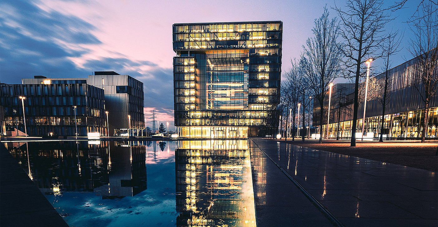 Moderne Immobilien im Ruhrgebiet wie das hier abgebildete thyssenkrupp Quartier in Essen. Zu sehen ist das beleuchtete Bürogebäude bei Abendhimmel mit Spiegelung im Wasser.