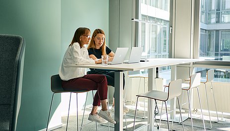 Two female employees are sitting at a meeting table looking at a laptop.