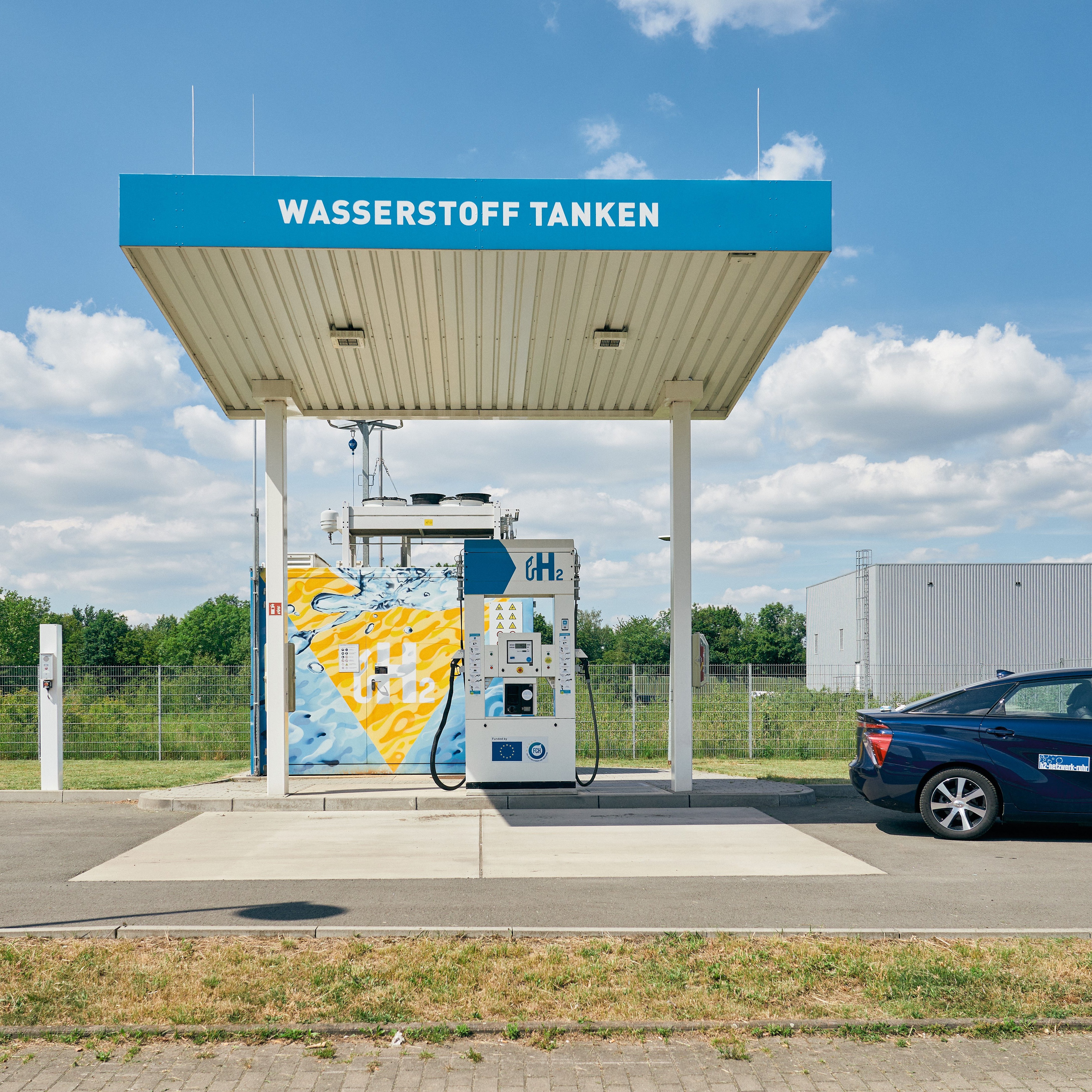 Abgebildet ist eine Wasserstofftankstelle auf der der Schriftzug Wasserstoff tanken abgebildet ist. Die Tankstelle wird von einem getanken Wasserstoffauto verlassen.