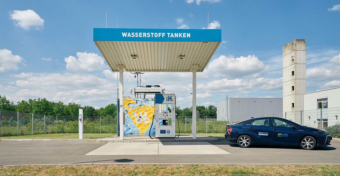 Abgebildet ist eine Wasserstofftankstelle auf der der Schriftzug Wasserstoff tanken abgebildet ist. Die Tankstelle wird von einem getanken Wasserstoffauto verlassen.